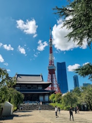 A traditional Japanese temple stands in the foreground, surrounded by lush greenery and several visitors walking around. In the background, a red and white broadcasting tower reaches into the bright blue sky, flanked by modern skyscrapers.