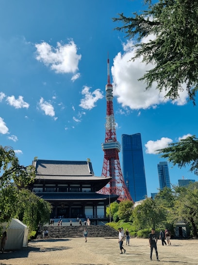 A traditional Japanese temple stands in the foreground, surrounded by lush greenery and several visitors walking around. In the background, a red and white broadcasting tower reaches into the bright blue sky, flanked by modern skyscrapers.
