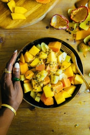 A bowl filled with various colorful, diced tropical fruits such as mango, papaya, and kiwi, topped with passion fruit seeds. A hand adorned with jewelry is resting on the bowl. The table also features a wooden cutting board with more diced fruits and some empty passion fruit shells, adding a rustic feel.