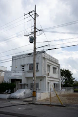 A two-story white house is situated beside a street, featuring a flat roof and several windows with security bars. A utility pole stands in the foreground with a complex network of electrical wires stretching across the scene. A car covered in a protective tarp is parked in the driveway. The house is bordered by a low white wall and there is a tree to the right in the background. The sky is overcast, adding a slightly muted quality to the scene.