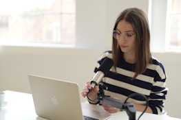 A person with long hair and glasses is seated at a white table, speaking into a microphone connected to a laptop. They are dressed in a striped shirt and are positioned in a well-lit room with large windows in the background.