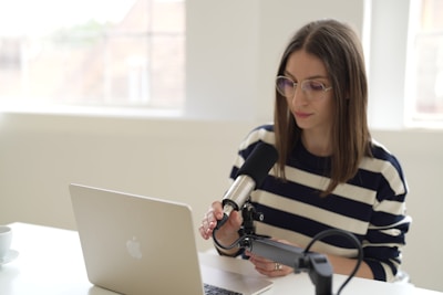 A person with long hair and glasses is seated at a white table, speaking into a microphone connected to a laptop. They are dressed in a striped shirt and are positioned in a well-lit room with large windows in the background.