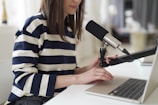 A woman wearing a striped sweater sits at a desk, speaking into a microphone attached to a boom arm. Her hands are resting on the keyboard of an open laptop, and she appears to be engaged in a podcast or recording session.