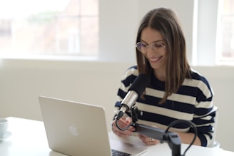 A person with long hair and glasses is seated at a white table, using a laptop. They are wearing a striped sweater and smiling while holding a phone. A microphone is positioned in front of them, suggesting a recording or broadcasting session. The environment is well-lit with a large window in the background.