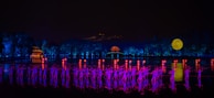 A nighttime scene features a group of performers illuminated in vibrant purple and pink lighting. They are positioned on a stage surrounded by water, with a backdrop of lush trees and a distant pavilion glowing in orange. A large, bright full moon is visible in the sky, adding to the dreamlike ambiance.