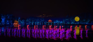 A nighttime scene features a group of performers illuminated in vibrant purple and pink lighting. They are positioned on a stage surrounded by water, with a backdrop of lush trees and a distant pavilion glowing in orange. A large, bright full moon is visible in the sky, adding to the dreamlike ambiance.