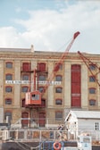 A historical industrial building with large arched windows and a prominent red crane attached to its facade. The structure is made of light-colored brick with red-painted accents, including doors and window frames. In the foreground, two people are standing near a small white building, and various industrial items are scattered around.
