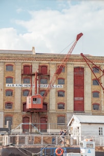 A historical industrial building with large arched windows and a prominent red crane attached to its facade. The structure is made of light-colored brick with red-painted accents, including doors and window frames. In the foreground, two people are standing near a small white building, and various industrial items are scattered around.