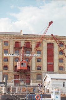 A historical industrial building with large arched windows and a prominent red crane attached to its facade. The structure is made of light-colored brick with red-painted accents, including doors and window frames. In the foreground, two people are standing near a small white building, and various industrial items are scattered around.