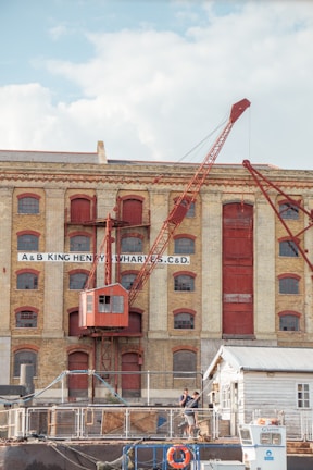 A historical industrial building with large arched windows and a prominent red crane attached to its facade. The structure is made of light-colored brick with red-painted accents, including doors and window frames. In the foreground, two people are standing near a small white building, and various industrial items are scattered around.