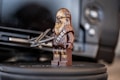 A LEGO figurine resembling a character with brown and beige details stands on a round, textured surface. It holds a small weapon and is positioned in front of a blurred background with metallic and black elements, likely a piece of camera equipment.