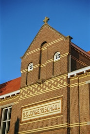 A brick building with a triangular gable roof, featuring decorative brickwork patterns. The building has a sign that reads 'R.K. Jongensschool' and includes arched windows with white frames. The sky is clear and blue, indicating a bright day.