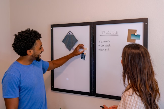 Two people are standing in front of a magnetic whiteboard with a to-do list. The person on the left, wearing a blue shirt, is pointing at the board, which has a square fabric sample and some color samples on it. The person on the right, with long hair, appears to be looking at the list, which includes tasks like groceries, car wash, and hair appointment.