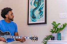 A man with short curly hair and glasses is playing an electric guitar. He is wearing a blue t-shirt and sitting indoors next to a framed abstract painting with swirling blue and green patterns. There is a potted plant on a white piece of furniture nearby and a switch plate on the wall.