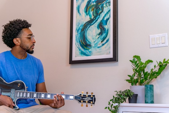 A man with short curly hair and glasses is playing an electric guitar. He is wearing a blue t-shirt and sitting indoors next to a framed abstract painting with swirling blue and green patterns. There is a potted plant on a white piece of furniture nearby and a switch plate on the wall.