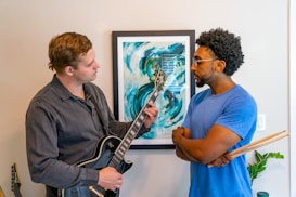 Two individuals are engaged in a musical conversation. One is holding a black electric guitar, playing or tuning it, while the other listens attentively, holding a pair of drumsticks. Behind them, a vibrant abstract painting adds a splash of color with its swirling blue and green tones. A plant is visible in the background, adding a touch of greenery to the room.