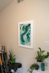 A room corner featuring a framed abstract painting dominated by swirling shades of blue and teal. Below the painting is a green houseplant placed on a white shelf, next to a green candle and a smaller black pot. To the left, a stool is situated beside several guitars leaning against the wall. The overall setting is neat and aesthetically arranged.