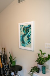 A room corner featuring a framed abstract painting dominated by swirling shades of blue and teal. Below the painting is a green houseplant placed on a white shelf, next to a green candle and a smaller black pot. To the left, a stool is situated beside several guitars leaning against the wall. The overall setting is neat and aesthetically arranged.
