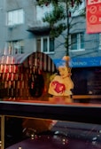 A whimsical artwork of a woman with blue curly hair holding a large red heart. The reflection in the glass combines with the background, showing a street scene with a gray building, windows, and some trees. Strung lights and a red banner add additional visual interest.