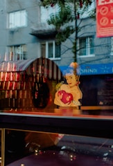 A whimsical artwork of a woman with blue curly hair holding a large red heart. The reflection in the glass combines with the background, showing a street scene with a gray building, windows, and some trees. Strung lights and a red banner add additional visual interest.