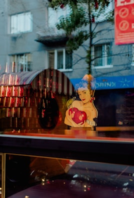 A whimsical artwork of a woman with blue curly hair holding a large red heart. The reflection in the glass combines with the background, showing a street scene with a gray building, windows, and some trees. Strung lights and a red banner add additional visual interest.