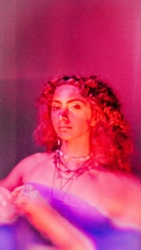 A person with curly hair is illuminated by dramatic, colorful lighting in shades of pink and purple, creating a vibrant and artistic atmosphere. The subject is wearing layered necklaces and a strapless top, with a contemplative expression on their face.