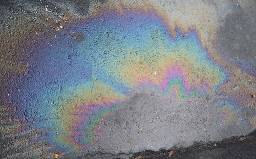 A vibrant, swirling pattern of iridescent colors covering the surface of a wet pavement. The colors range through shades of blue, purple, green, yellow, and pink, creating an eye-catching contrast against the dark and rough texture of the asphalt.