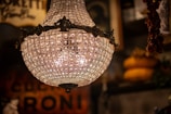 A vintage, ornate chandelier with numerous crystals is prominently featured, creating an intricate pattern of light. The background is blurred, highlighting warm tones and hints of rustic decor, possibly from an antique shop or an old interior setting.