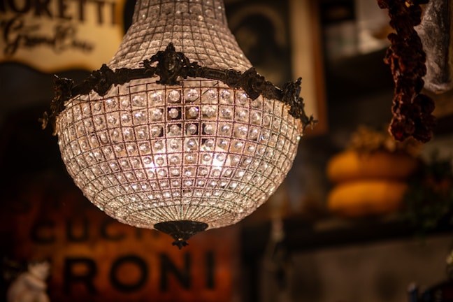 A vintage, ornate chandelier with numerous crystals is prominently featured, creating an intricate pattern of light. The background is blurred, highlighting warm tones and hints of rustic decor, possibly from an antique shop or an old interior setting.