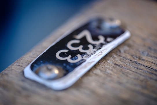 A close-up of a metal plate with the number '333' embossed on it, attached to a wooden surface. The plate appears slightly worn with visible scratches and two rivets securing it to the surface.