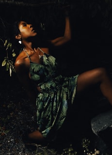 A woman in a tropical dress poses dramatically in a dimly lit environment, with shadows highlighting her facial profile and the leaves around her blending into the darkness.