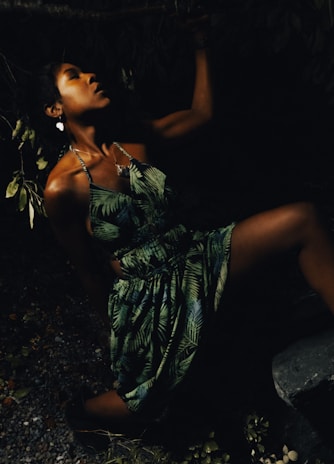 A woman in a tropical dress poses dramatically in a dimly lit environment, with shadows highlighting her facial profile and the leaves around her blending into the darkness.