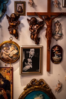 A wall display featuring various religious and classical art pieces. Several angel and cherub sculptures are mounted, highlighting ornate details and various poses. A wooden crucifix is prominently displayed, alongside framed paintings depicting religious and historical figures. The arrangement is eclectic, blending different artistic styles.