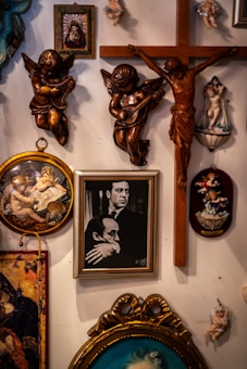 A wall display featuring various religious and classical art pieces. Several angel and cherub sculptures are mounted, highlighting ornate details and various poses. A wooden crucifix is prominently displayed, alongside framed paintings depicting religious and historical figures. The arrangement is eclectic, blending different artistic styles.