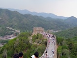 A section of the Great Wall of China stretches across a hilly landscape with lush green vegetation. Numerous people walk along the wall, enjoying the scenic view. The surrounding mountains are covered in greenery, creating a serene backdrop.
