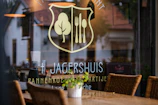 A restaurant window displays a logo with a fork and knife inside a shield, alongside a tree symbol. The name 'Jagershuis' and other text are visible. Inside, wicker chairs and tables are set up, while reflections of the surrounding area are seen on the glass.