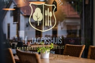 A restaurant window displays a logo with a fork and knife inside a shield, alongside a tree symbol. The name 'Jagershuis' and other text are visible. Inside, wicker chairs and tables are set up, while reflections of the surrounding area are seen on the glass.