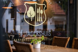 A restaurant window displays a logo with a fork and knife inside a shield, alongside a tree symbol. The name 'Jagershuis' and other text are visible. Inside, wicker chairs and tables are set up, while reflections of the surrounding area are seen on the glass.