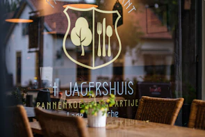 A restaurant window displays a logo with a fork and knife inside a shield, alongside a tree symbol. The name 'Jagershuis' and other text are visible. Inside, wicker chairs and tables are set up, while reflections of the surrounding area are seen on the glass.