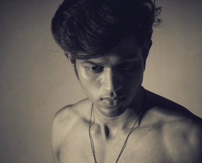 A young man with a serious expression is captured in a monochromatic photograph. The light casts shadows on his bare shoulders and chest, highlighting the contours of his face and hair. He appears introspective, looking slightly downwards.