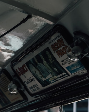 An interior view of a retro vehicle featuring overhead advertisements. The advertisement displays stylized artwork and bold text, some of which reads 'Factory Prices' and highlights a luxury ship. The subdued lighting creates a vintage ambiance with glossy metal surfaces.