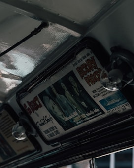 An interior view of a retro vehicle featuring overhead advertisements. The advertisement displays stylized artwork and bold text, some of which reads 'Factory Prices' and highlights a luxury ship. The subdued lighting creates a vintage ambiance with glossy metal surfaces.