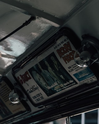 An interior view of a retro vehicle featuring overhead advertisements. The advertisement displays stylized artwork and bold text, some of which reads 'Factory Prices' and highlights a luxury ship. The subdued lighting creates a vintage ambiance with glossy metal surfaces.