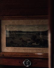 A vintage photograph is displayed within a wooden frame. The image features a pastoral landscape with rolling hills, scattered trees, and a few small structures. The scene is labeled as 'Wingham, N.S.W.' with additional text describing the Manning River area. The setup includes a metallic reserved sign with the number '9' at the bottom of the frame.