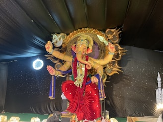 A large and intricately designed statue of a deity, with multiple arms holding various symbolic items. The figure is adorned with elaborate headgear and jewelry, standing against a backdrop of dark fabric with subtle lighting accents.