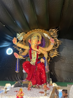 A large, intricately decorated statue of a deity with an elephant head, multiple arms, and adorned with ornate jewelry and garments in vibrant colors. The statue is set against a dark, star-like backdrop and surrounded by smaller religious icons and offerings.