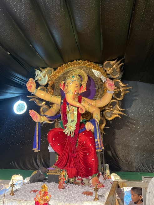 A large, intricately decorated statue of a deity with an elephant head, multiple arms, and adorned with ornate jewelry and garments in vibrant colors. The statue is set against a dark, star-like backdrop and surrounded by smaller religious icons and offerings.