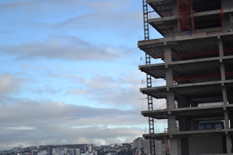 A multi-story building under construction stands prominently on the right side of the image. The structure is made of concrete with visible floors and metal scaffolding. In the background, a cityscape with numerous low-rise buildings extends across the horizon under a partly cloudy sky.