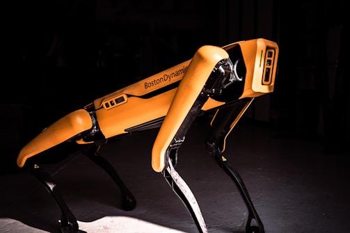 A robotic dog is highlighted against a dark background. The robot has a yellow body with four black legs and is marked with the name Boston Dynamics. The lighting creates a dramatic contrast, focusing attention on the machine's design and structure.