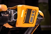 A detailed view of a robotic device featuring a bright orange casing with multiple cameras or sensors. The design includes perforated sections for ventilation and exposed mechanical parts, indicating advanced technology and engineering.